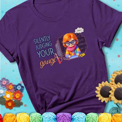 Purple t-shirt with a serious-looking crocheter in rainbow clothing and big glasses, holding a tape measure under the phrase “Silently Judging Your Gauge.”