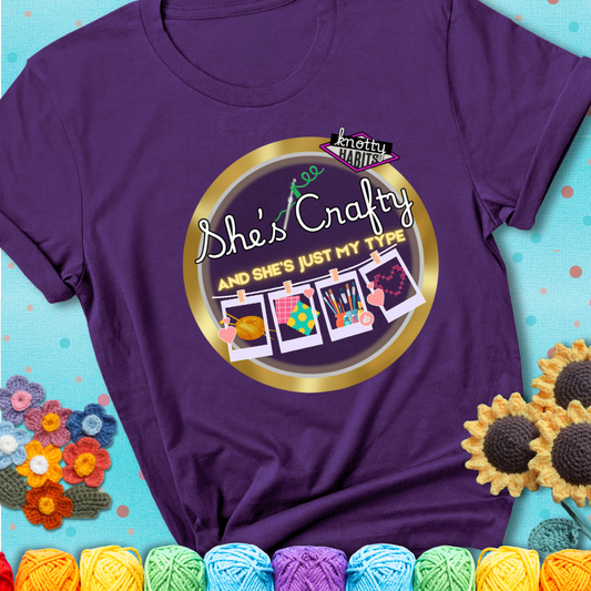 Text on purple tee reads "She’s Crafty & She’s Just My Type" with yarn, fabric, painting, and cross-stitch icons hanging like Polaroids