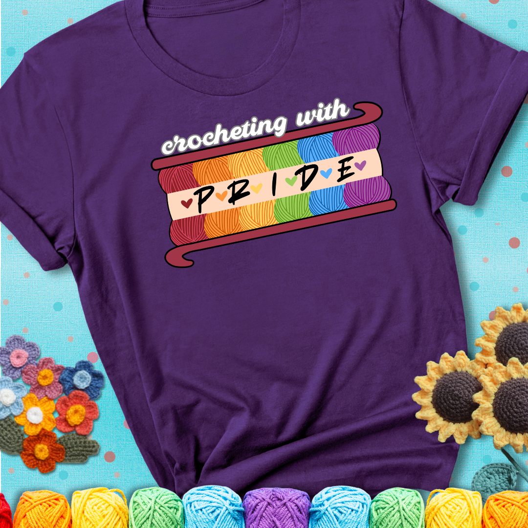 Yarn skeins in rainbow colors stacked in a hook shape. Text reads “Crocheting With Pride” showing a purple tee with a teal sunflower background