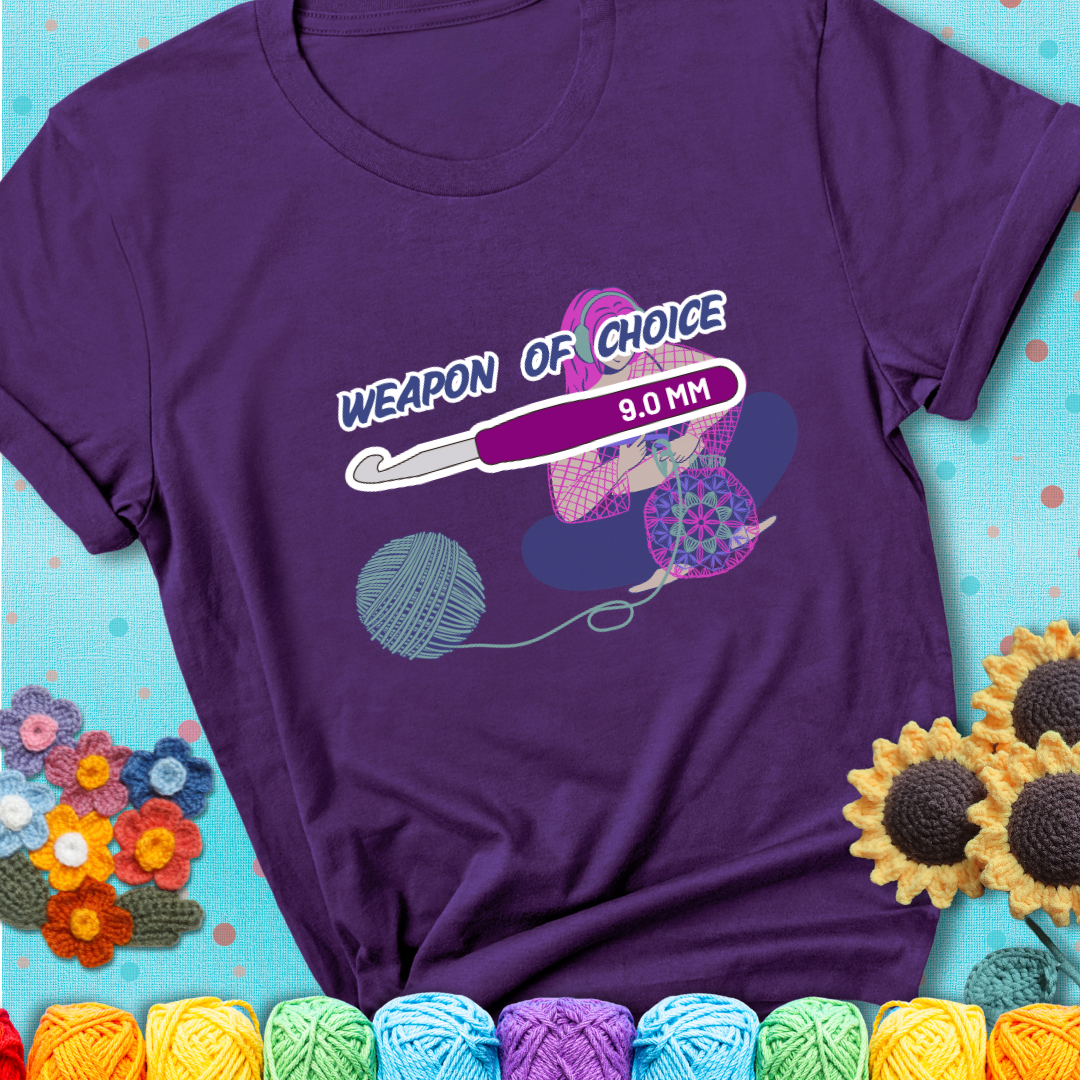 Purple t-shirt with an Illustration of a crocheter sitting cross-legged with a 9.0 mm crochet hook above her, labeled “Weapon of Choice.” She’s crocheting a colorful mandala-style project from a large yarn ball.