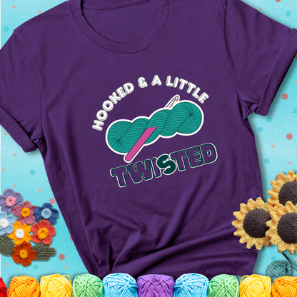 A teal skein of yarn with a hot pink crochet hook through it, captioned “Hooked & a Little Twisted” on a purple t-shirt.