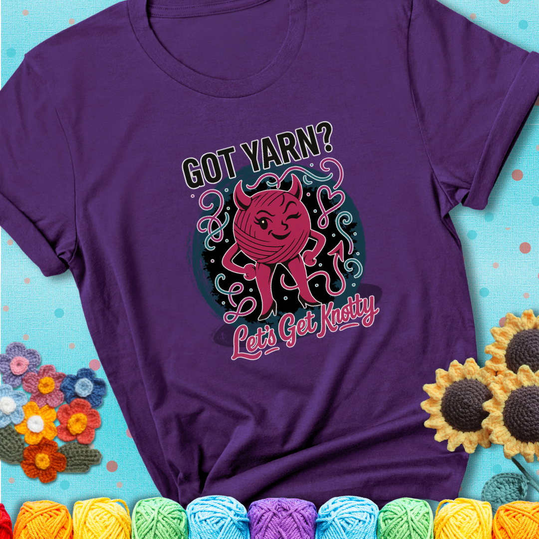 Purple t-shirt with a cheeky pink yarn ball wearing devil horns, arms, and legs, winking beneath the phrase “Got Yarn? Let’s Get Knotty