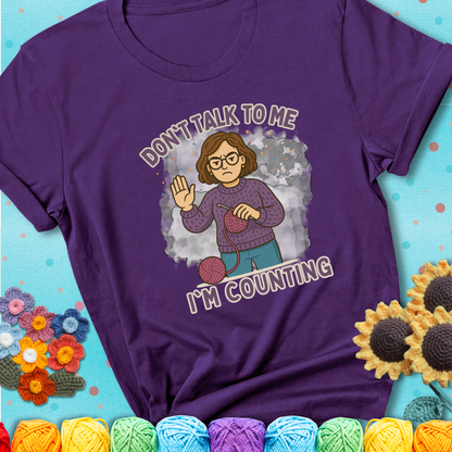 Purple t-shirt with an annoyed maker holding up a hand beside yarn, with the phrase “Don’t Talk To Me, I’m Counting.”