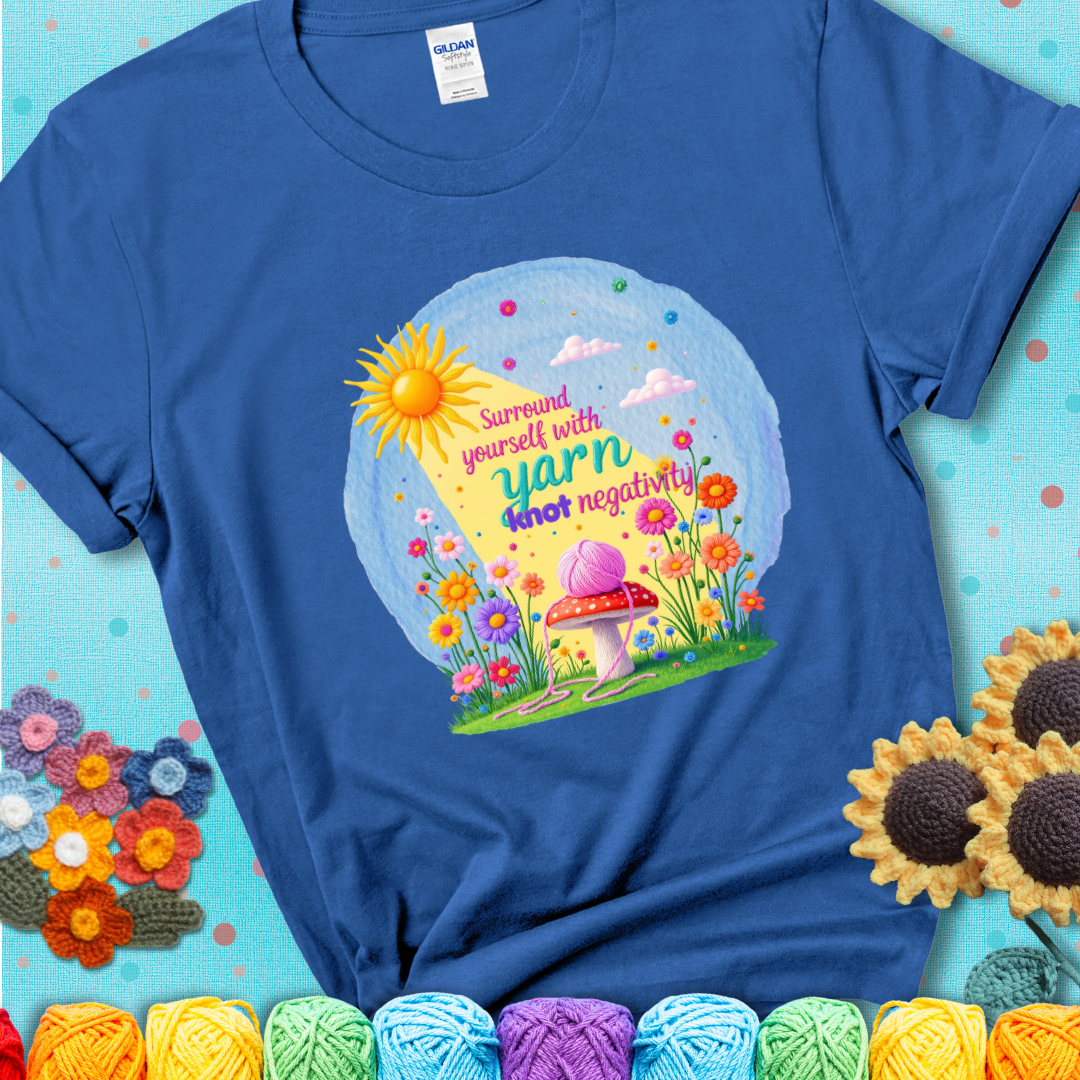 Blue t-shirt featuring a happy spring garden scene with a sunbeam, flowers, and a pink yarn ball on a mushroom, with the quote "Surround yourself with yarn, knot negativity."