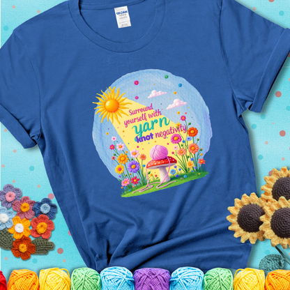 Blue t-shirt featuring a happy spring garden scene with a sunbeam, flowers, and a pink yarn ball on a mushroom, with the quote "Surround yourself with yarn, knot negativity."