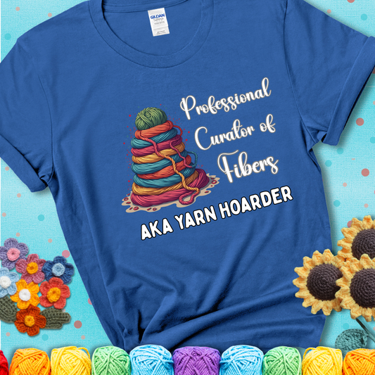 A blue t-shirt showing a pyramid of rainbow yarn topped with green ball; text reads “Professional Curator of Fibers AKA Yarn Hoarder”, a playful shirt for serious stashers.