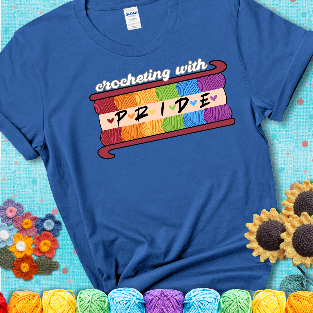 Yarn skeins in rainbow colors stacked in a hook shape. Text reads “Crocheting With Pride” showing a blue tee with a teal sunflower background