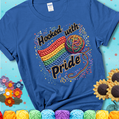 A rainbow-striped crochet swatch paired with a multicolor yarn ball and bold “Hooked With Pride” lettering. Confetti surrounds the vibrant scene showing a blue tee with a teal sunflower background