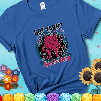 Royal blue t-shirt with a cheeky pink yarn ball wearing devil horns, arms, and legs, winking beneath the phrase “Got Yarn? Let’s Get Knotty