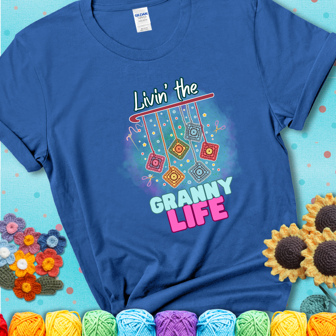Blue t-shirt shows colorful crochet granny squares hanging from a hook with text “Livin’ the Granny Life” in bold pink and blue font; a fun nod to classic crochet.