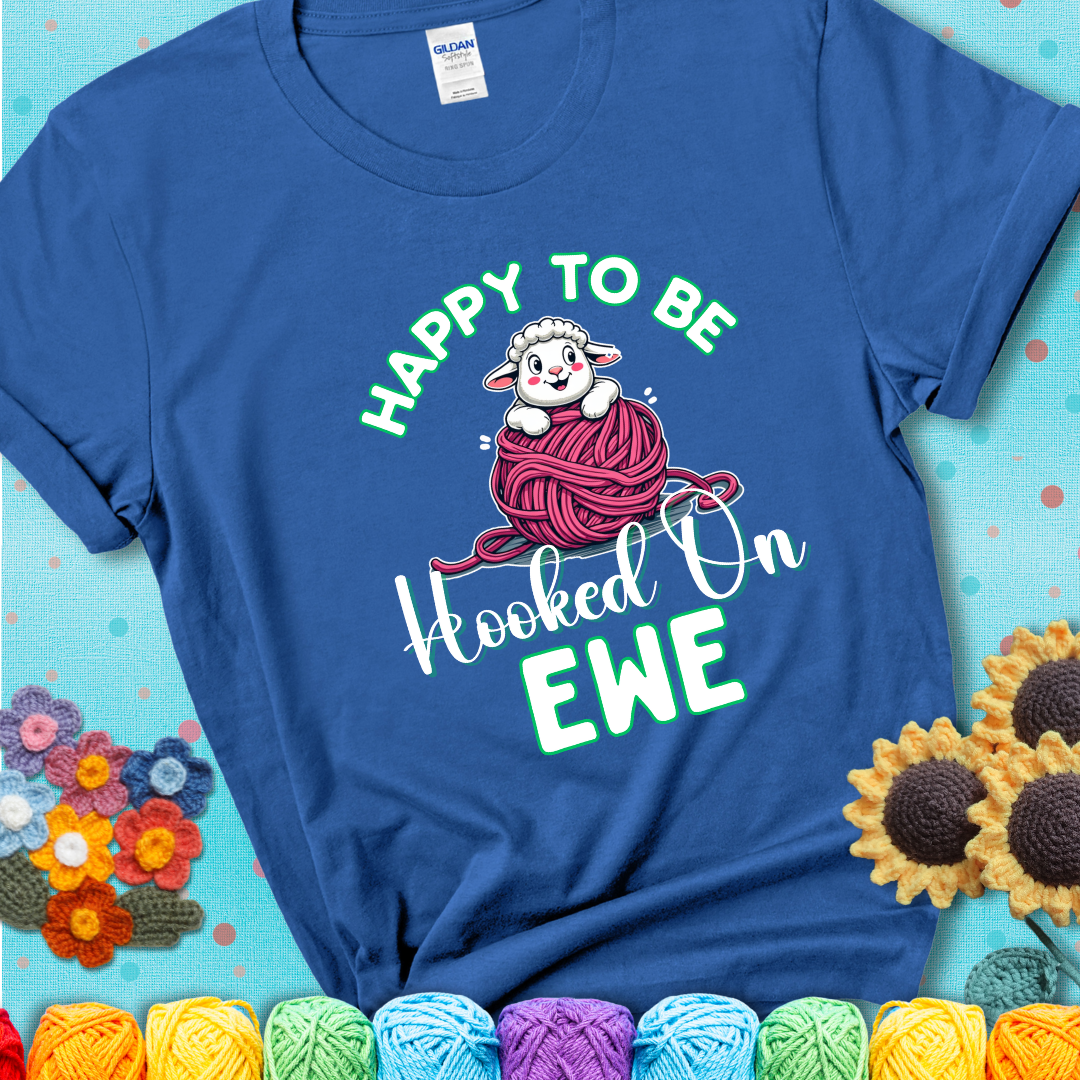 Blue t-shirt with smiling cartoon sheep peeks from a pink yarn ball, surrounded by green text “Happy to Be Hooked on Ewe”, a playful and affectionate sheep pun for crocheters.