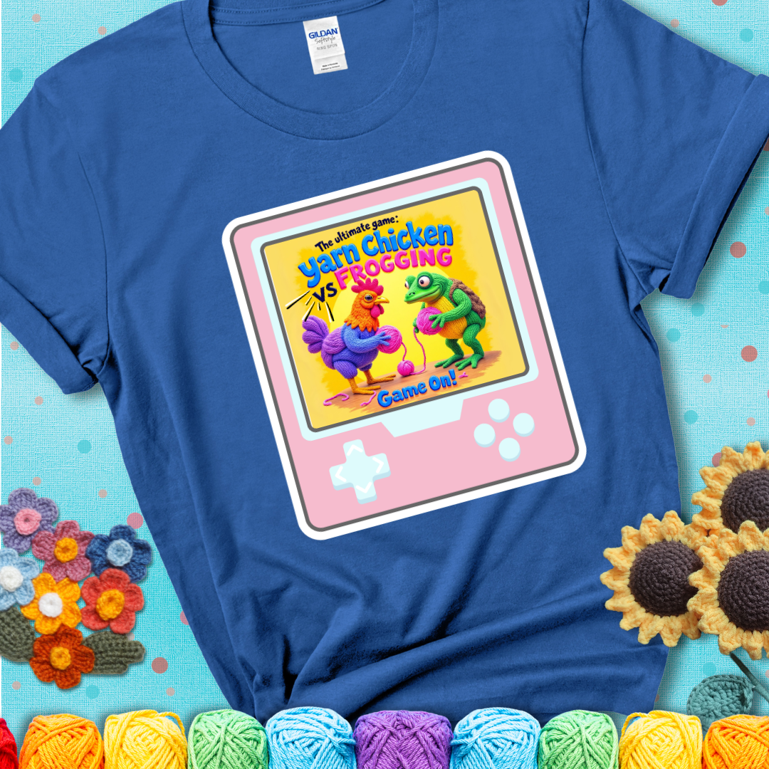 A blue t-shirt featuring a cartoon yarn chicken and frog face off with text “Yarn Chicken vs Frogging: Game On!” on a retro, handheld video game meets crochet humor battle design.