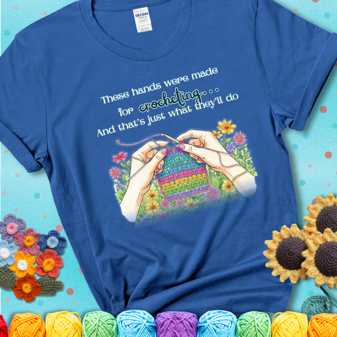 Royal blue t-shirt with an illustration of two hands crocheting a rainbow swatch, surrounded by colorful flowers and the phrase “These hands were made for crocheting… and that’s just what they’ll do.”