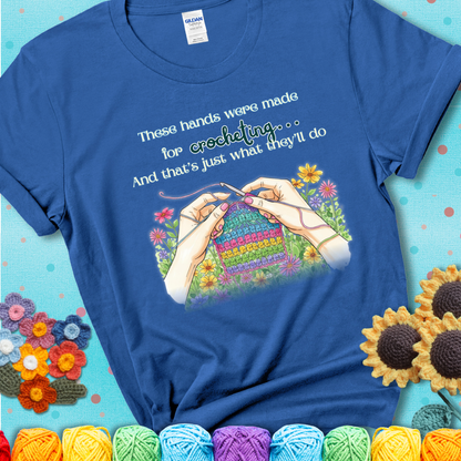 Royal blue t-shirt with an illustration of two hands crocheting a rainbow swatch, surrounded by colorful flowers and the phrase “These hands were made for crocheting… and that’s just what they’ll do.”