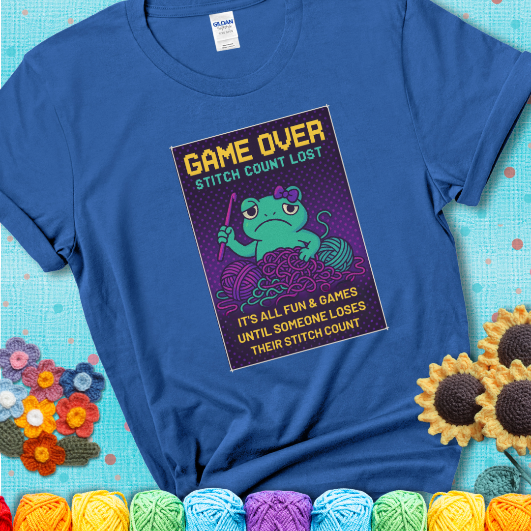 Blue t-shirt shows a pixel-style design of a grumpy yarn frog holding a hook, surrounded by yarn chaos, with the text “Game Over. Stitch Count Lost.”