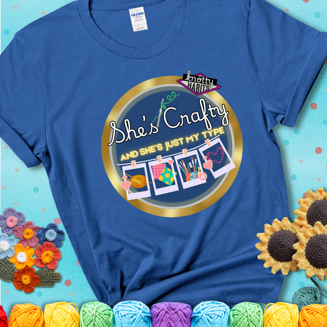 Text on royal blue tee reads "She’s Crafty & She’s Just My Type" with yarn, fabric, painting, and cross-stitch icons hanging like Polaroids