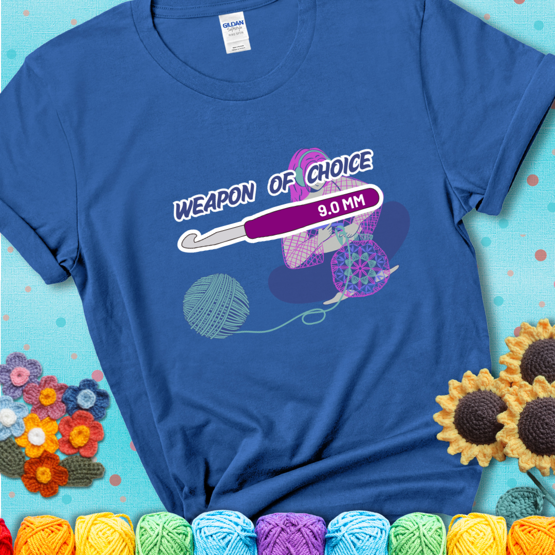 Blue t-shirt with an Illustration of a crocheter sitting cross-legged with a 9.0 mm crochet hook above her, labeled “Weapon of Choice.” She’s crocheting a colorful mandala-style project from a large yarn ball.