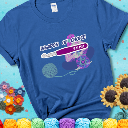 Blue t-shirt with an Illustration of a crocheter sitting cross-legged with a 9.0 mm crochet hook above her, labeled “Weapon of Choice.” She’s crocheting a colorful mandala-style project from a large yarn ball.