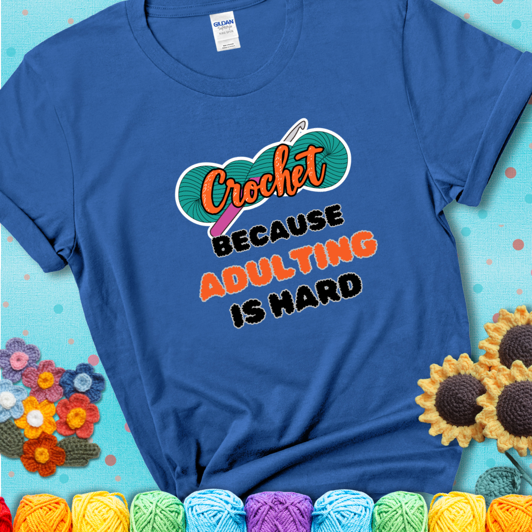 Blue t-shirt featuring a bold design with the phrase “Crochet Because Adulting Is Hard” featuring a green skein of yarn with a pink crochet hook. The word “Crochet” is in a script font, while “Adulting is hard” is in chunky textured letters.