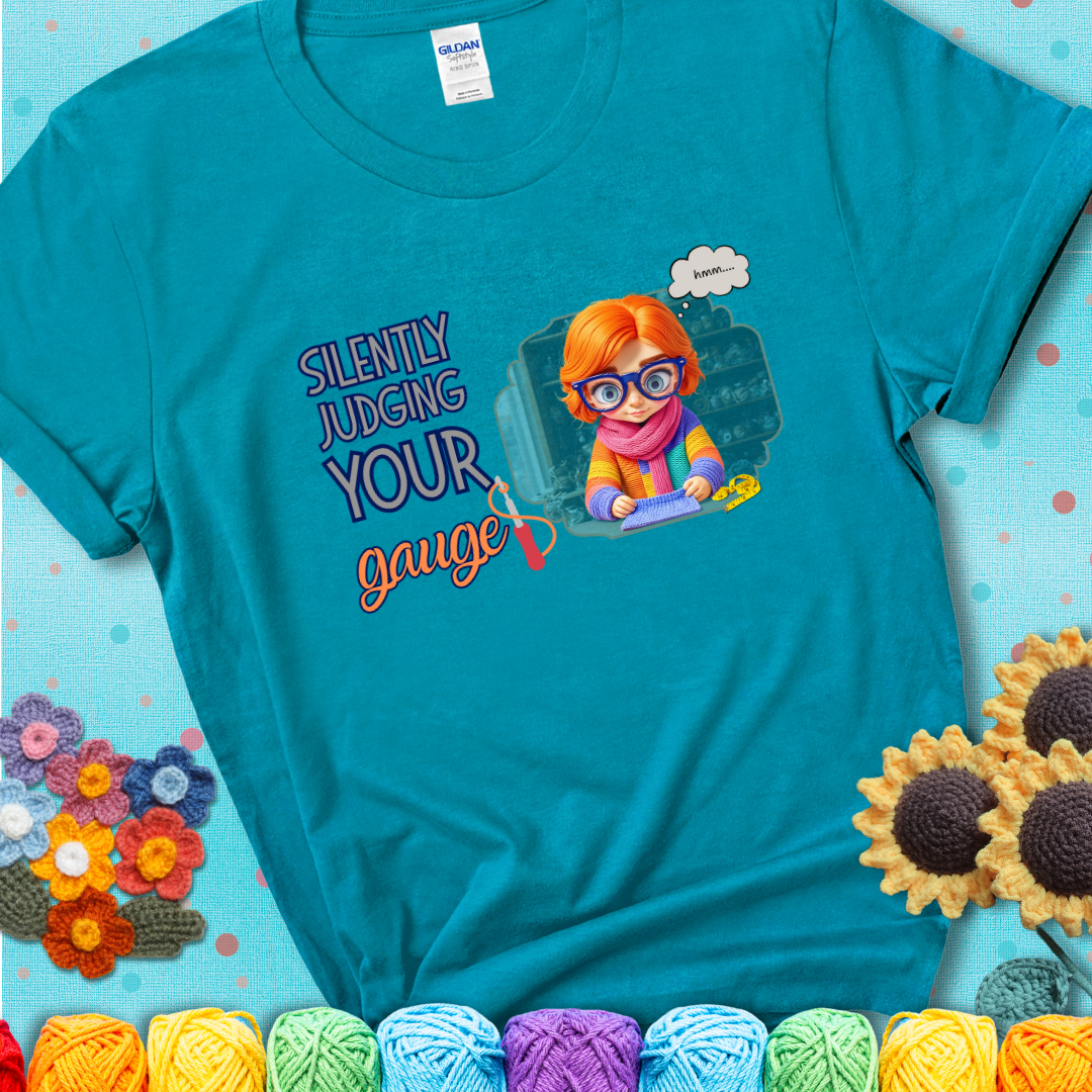Tropical Blue t-shirt with a serious-looking crocheter in rainbow clothing and big glasses, holding a tape measure under the phrase “Silently Judging Your Gauge.”