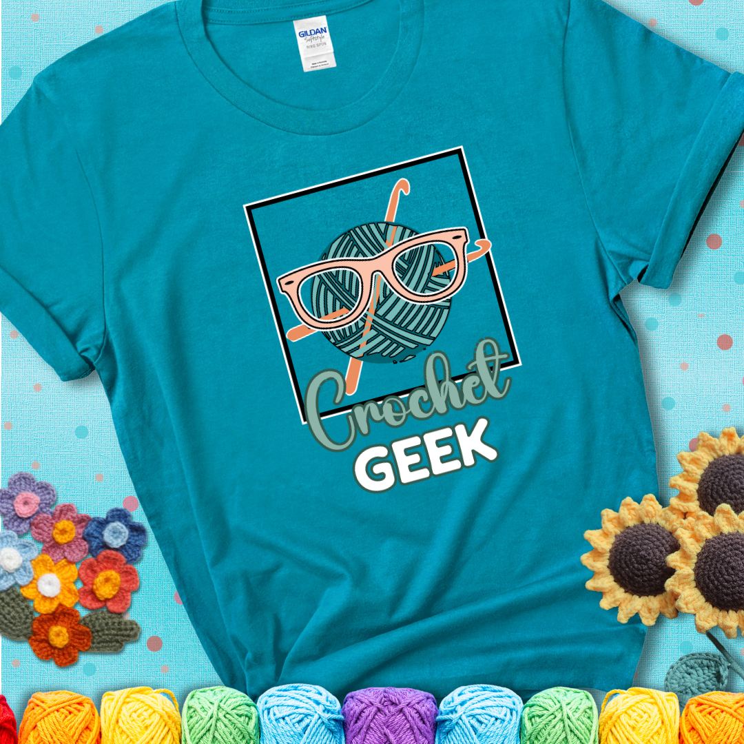 A teal yarn ball with crisscrossed hooks wears oversized nerdy glasses with the phrase “Crochet Geek” in bold, fun text below on a teal t-shirt.