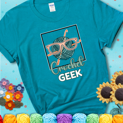 A teal yarn ball with crisscrossed hooks wears oversized nerdy glasses with the phrase “Crochet Geek” in bold, fun text below on a teal t-shirt.
