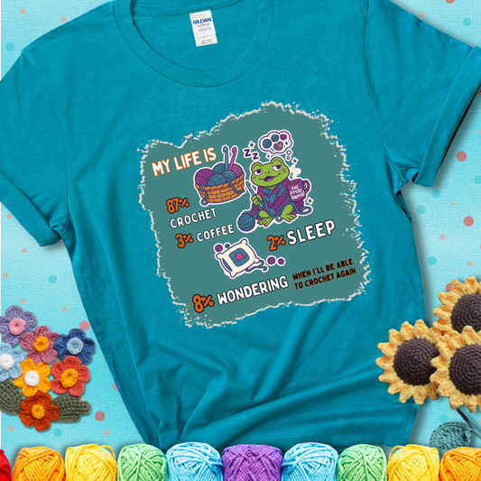 Tropical blue t-shirt with a crocheting frog, coffee, yarn, and text showing a playful life breakdown: 87% crochet, 3% coffee, 2% sleep, 8% wondering when to crochet again.