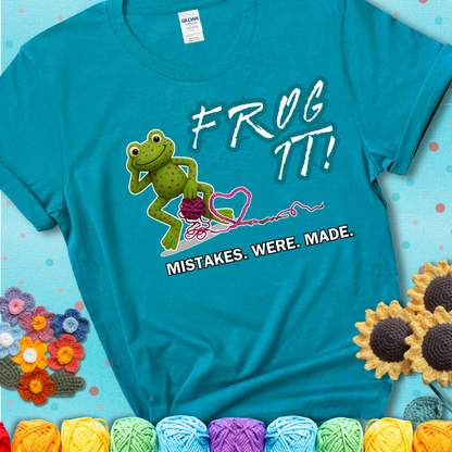 Green frog with yarn and heart-shaped loop. Text reads: “Frog It! Mistakes Were Made.” A playful crochet tee for anyone who's ever had to rip it back, showing a blue-green teal tee with a teal sunflower background