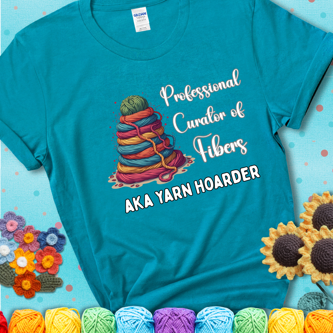 A teal t-shirt showing a pyramid of rainbow yarn topped with green ball; text reads “Professional Curator of Fibers AKA Yarn Hoarder”, a playful shirt for serious stashers.