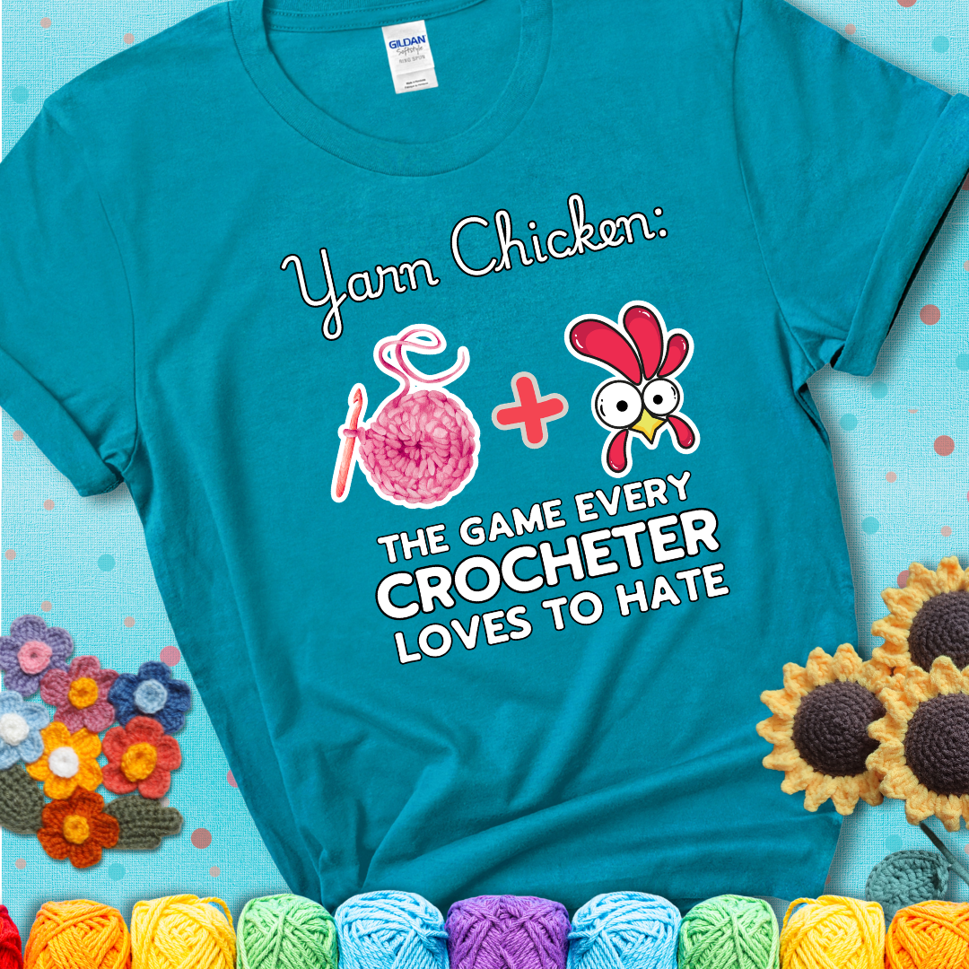 Cartoon chicken inside a yarn ball with crochet circle background. Text reads: “Yarn Chicken: The Game Every Crocheter Loves to Lose.” A fun and relatable tee for fiber artists who like to live on the edge, showing a blue-green teal tee with an aqua sunflower background