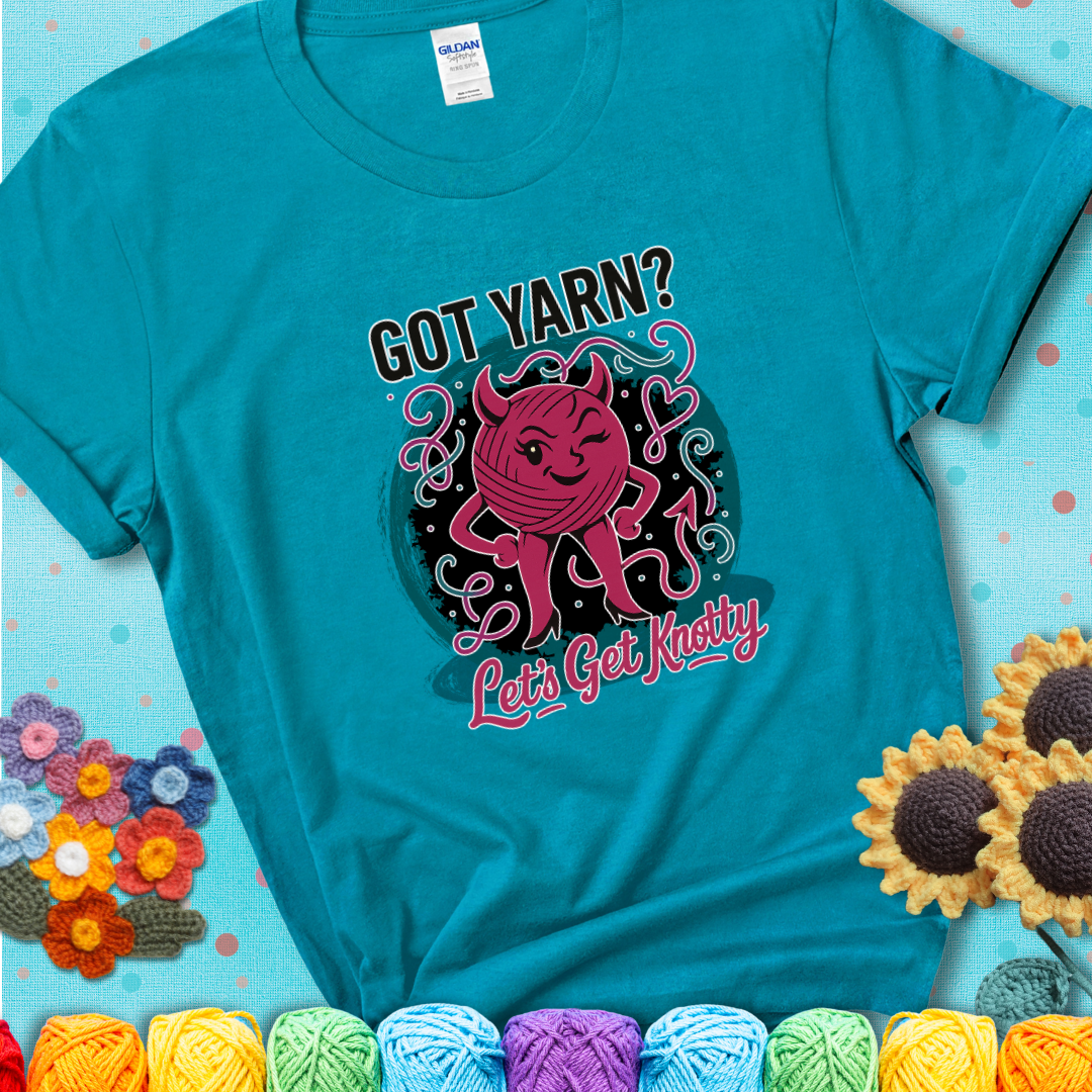 Tropical blue t-shirt with a cheeky pink yarn ball wearing devil horns, arms, and legs, winking beneath the phrase “Got Yarn? Let’s Get Knotty