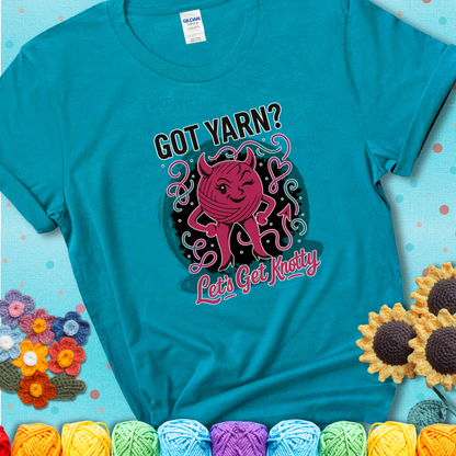 Tropical blue t-shirt with a cheeky pink yarn ball wearing devil horns, arms, and legs, winking beneath the phrase “Got Yarn? Let’s Get Knotty
