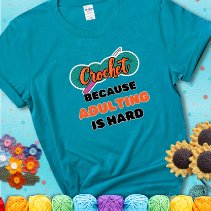 Teal t-shirt featuring a bold design with the phrase “Crochet Because Adulting Is Hard” featuring a green skein of yarn with a pink crochet hook. The word “Crochet” is in a script font, while “Adulting is hard” is in chunky textured letters.