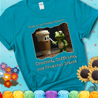 A crocheted frog holding a crochet hook confronts a surprised coffee cup in a dim alleyway with text: "Next up on Knotty Hooker: Crochet, Caffeine, and Criminal Intent," showing a blue-green teal tee with a teal sunflower background