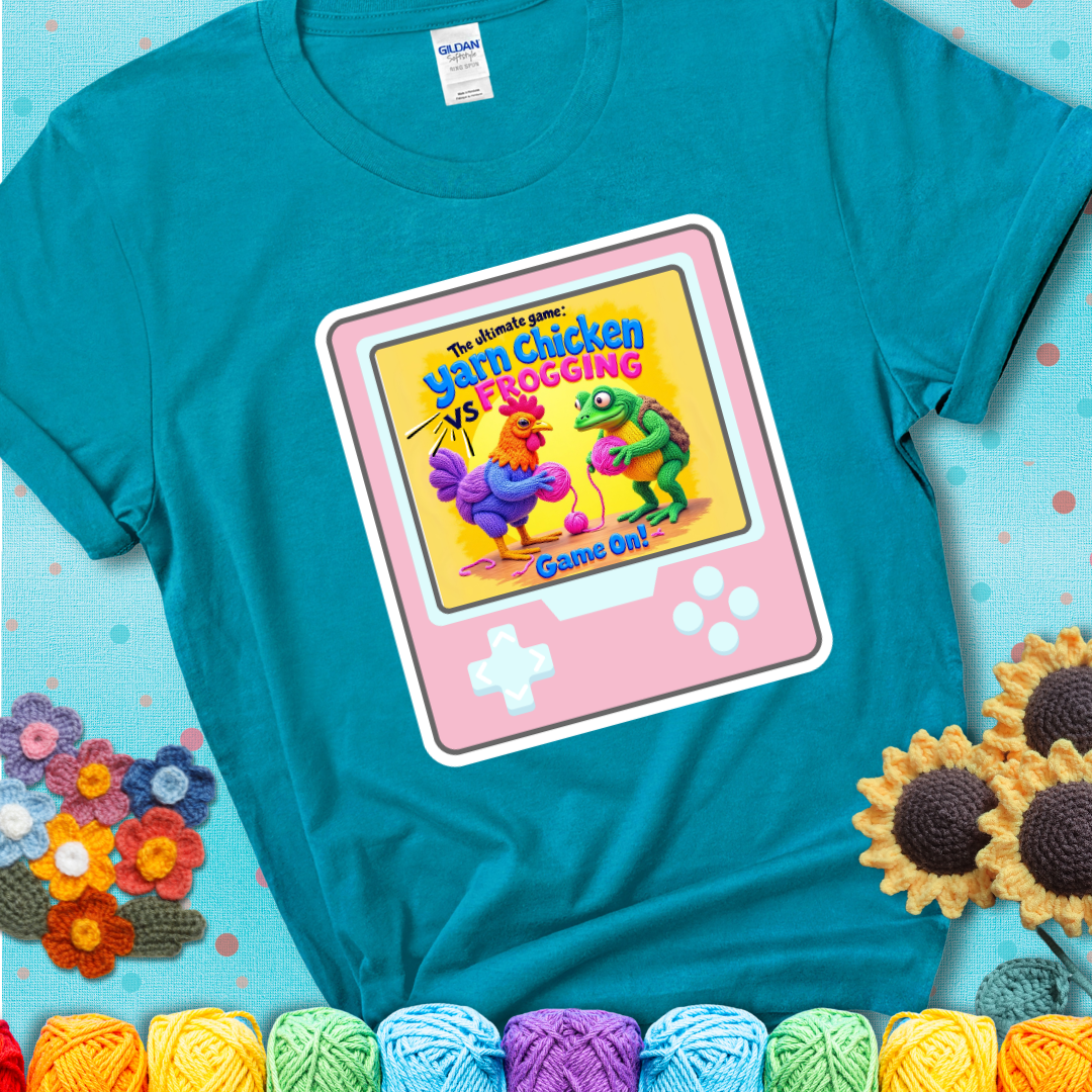 A teal t-shirt featuring a cartoon yarn chicken and frog face off with text “Yarn Chicken vs Frogging: Game On!” on a retro, handheld video game meets crochet humor battle design.