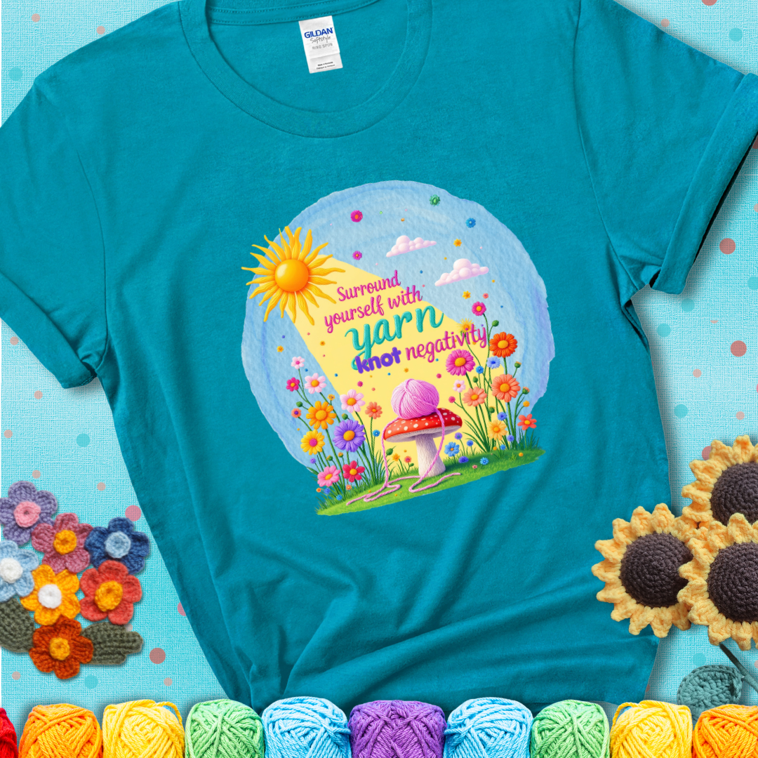 Teal t-shirt featuring a happy spring garden scene with a sunbeam, flowers, and a pink yarn ball on a mushroom, with the quote "Surround yourself with yarn, knot negativity."