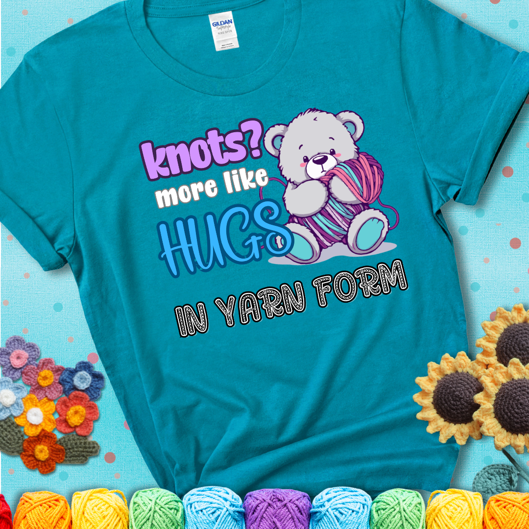 Cute teddy bear holding colorful yarn ball with hearts. Text reads: “Knots? More Like Hugs in Yarn Form.” Perfect for cozy gift-givers and yarn lovers, showing a blue-green teal tee with an aqua sunflower background