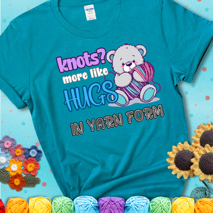 Cute teddy bear holding colorful yarn ball with hearts. Text reads: “Knots? More Like Hugs in Yarn Form.” Perfect for cozy gift-givers and yarn lovers, showing a blue-green teal tee with an aqua sunflower background