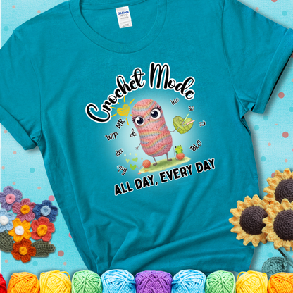 Teal t-shirt with a kawaii-style rainbow yarn character surrounded by crochet stitch abbreviations and holding a yarn heart, with text “Crochet Mode All Day, Every Day.”