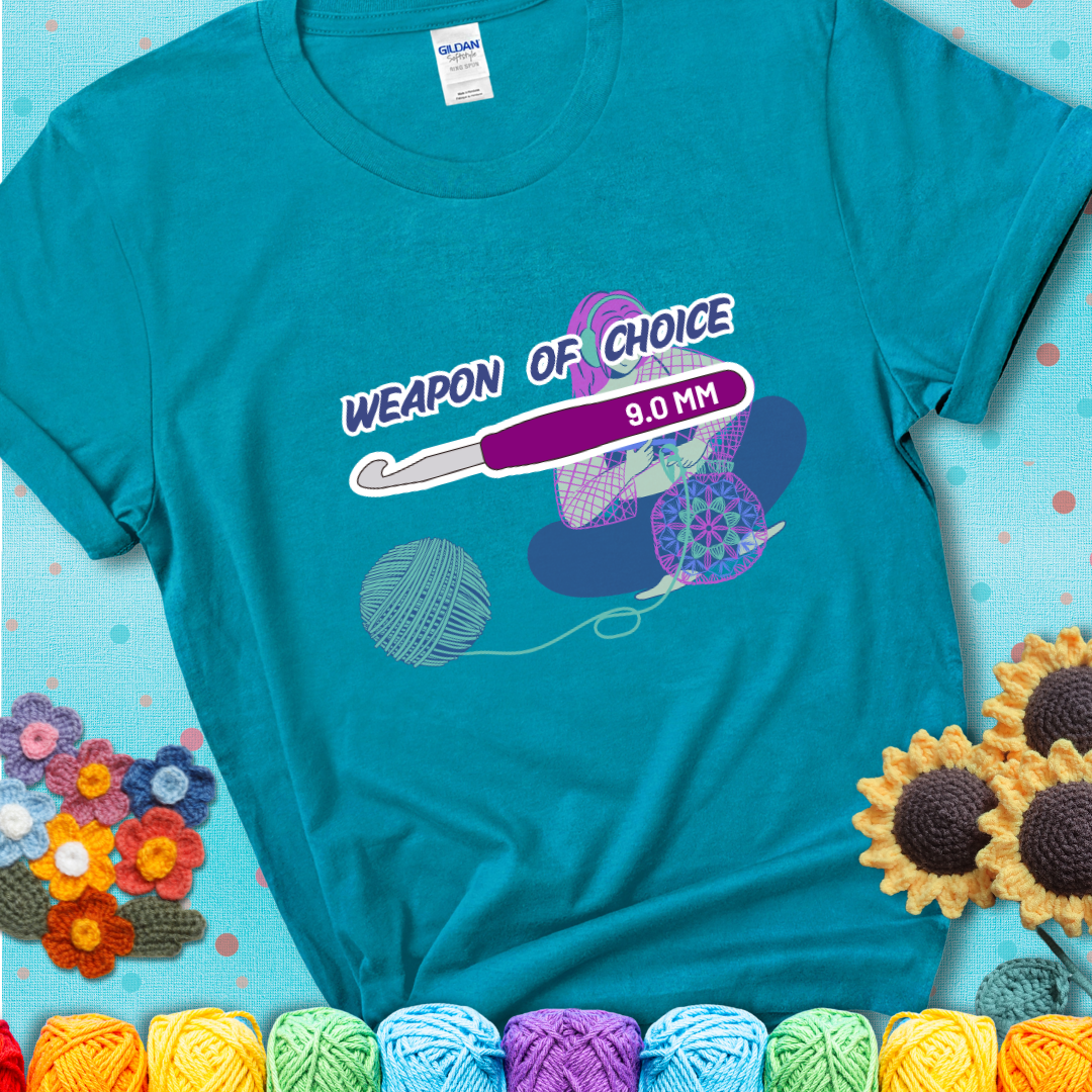 Teal t-shirt with an Illustration of a crocheter sitting cross-legged with a 9.0 mm crochet hook above her, labeled “Weapon of Choice.” She’s crocheting a colorful mandala-style project from a large yarn ball.