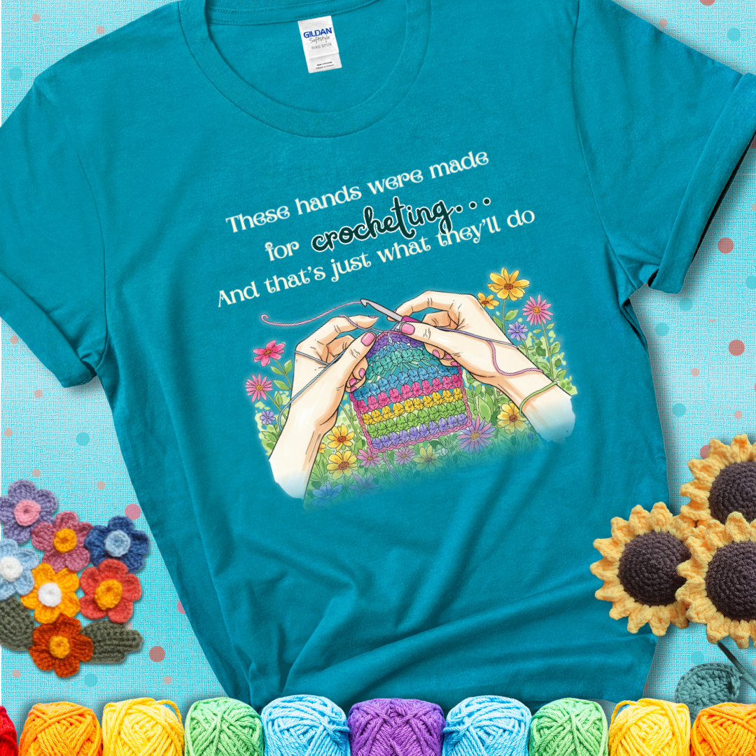 Tropical blue t-shirt with an illustration of two hands crocheting a rainbow swatch, surrounded by colorful flowers and the phrase “These hands were made for crocheting… and that’s just what they’ll do.”