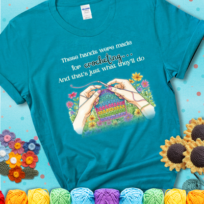 Tropical blue t-shirt with an illustration of two hands crocheting a rainbow swatch, surrounded by colorful flowers and the phrase “These hands were made for crocheting… and that’s just what they’ll do.”