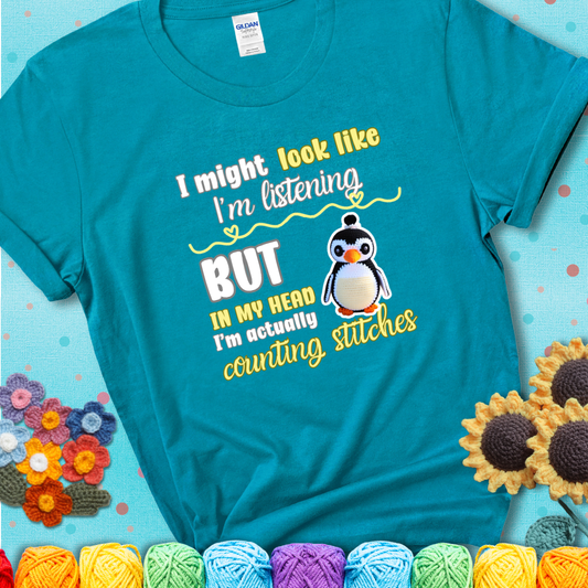 Teal t-shirt shows a crocheted penguin next to the text “I might look like I’m listening but in my head I’m actually counting stitches.”