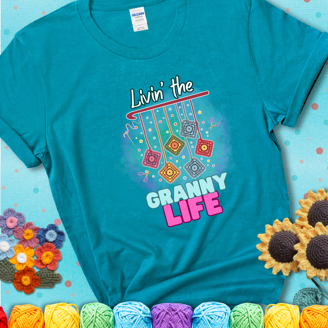 Teal t-shirt shows colorful crochet granny squares hanging from a hook with text “Livin’ the Granny Life” in bold pink and blue font; a fun nod to classic crochet.