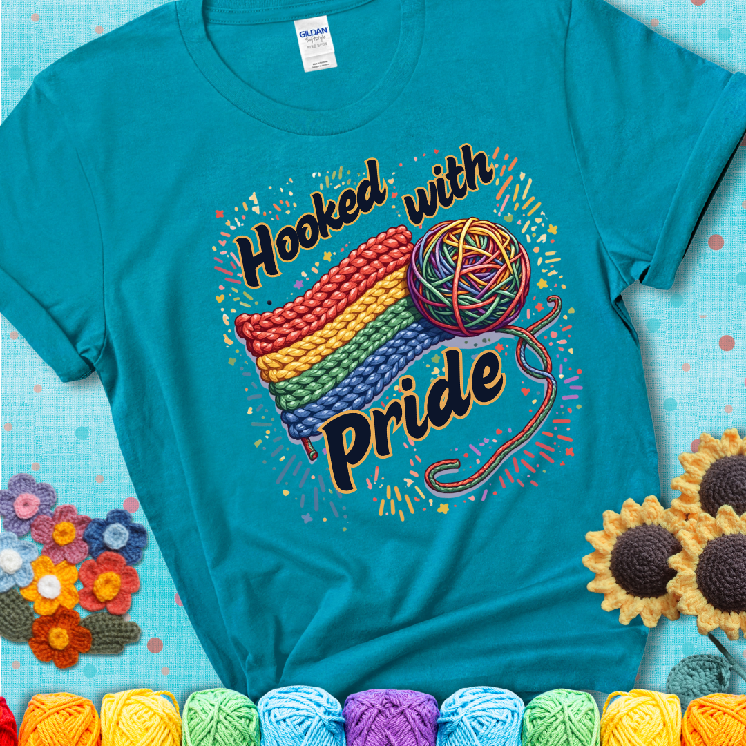 A rainbow-striped crochet swatch paired with a multicolor yarn ball and bold “Hooked With Pride” lettering. Confetti surrounds the vibrant scene showing a blue-green teal tee with an aqua  sunflower background