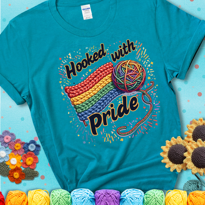 A rainbow-striped crochet swatch paired with a multicolor yarn ball and bold “Hooked With Pride” lettering. Confetti surrounds the vibrant scene showing a blue-green teal tee with an aqua  sunflower background