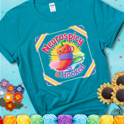 Teal t-shirt featuring a smiling red chili pepper wrapped in yarn with a crochet hook, text says “Neurospicy & Hooked.”