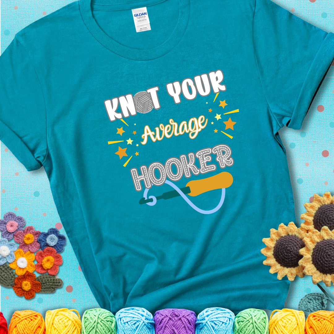Bold lettering on a teal t-shirt with a yarn hook graphic. Text reads: “I'm Not Your Average Hooker.” A cheeky crochet tee for confident crafters who own their skills.