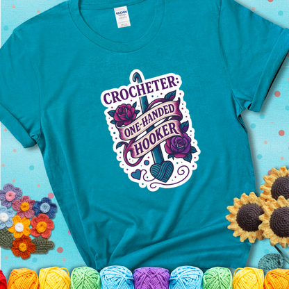 Tropical Blue t-shirt with a tattoo-style crochet design featuring a crochet hook, roses, and the phrase “Crocheter: One-Handed Hooker.”