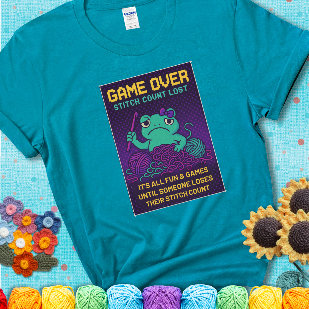 Teal t-shirt shows a pixel-style design of a grumpy yarn frog holding a hook, surrounded by yarn chaos, with the text “Game Over. Stitch Count Lost.”